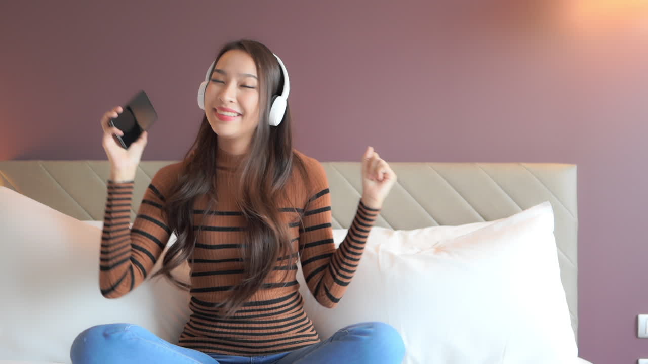 While sitting on a big comfortable bed, young attractive woman begins to dance with the music through her smartphone and headphones