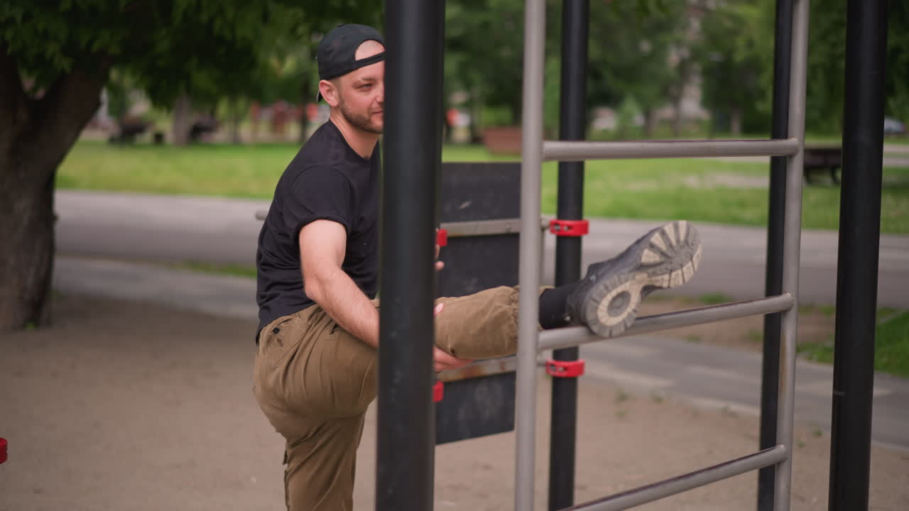 Hombre blanco estirando la pierna en una barra, calentamiento de calistenia en un parque urbano con expresión sonriente, gorra y pantalones casuales, barras de metal y árboles verdes de fondo, rutina de movilidad de los isquiotibiales, entrenamiento individual.