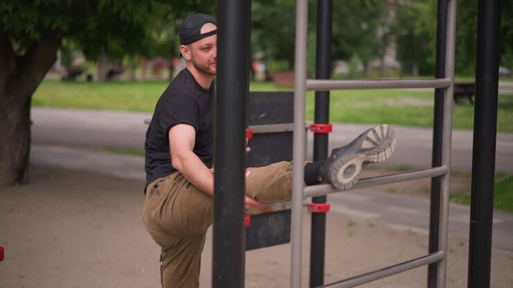 white man stretching leg on bar, urban park calisthenics warmup with smiling expression, casual cap and pants, metal bars and green trees background, hamstring mobility routine, solo training
