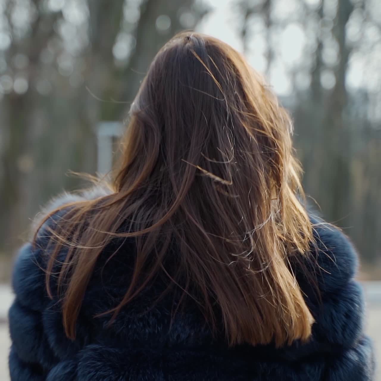 Beautiful woman with a plastic cup outdoors. Pretty girl in fur coat walking in the park. Back view of a brunette in the park. Slow motion.