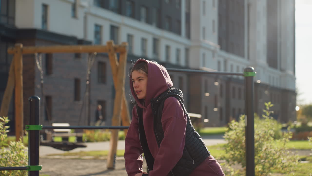 Outdoor sportswoman in hoodie and quilted vest leans into deep lunge gripping iron bar for support while other people move in urban park under warm sunset light showing focused relief on her face