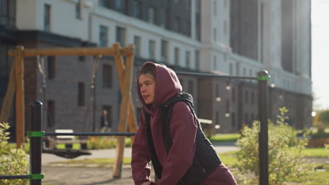 outdoor sportswoman in hoodie and quilted vest leans into deep lunge gripping iron bar for support while other people move in urban park under warm sunset light showing focused relief on her face