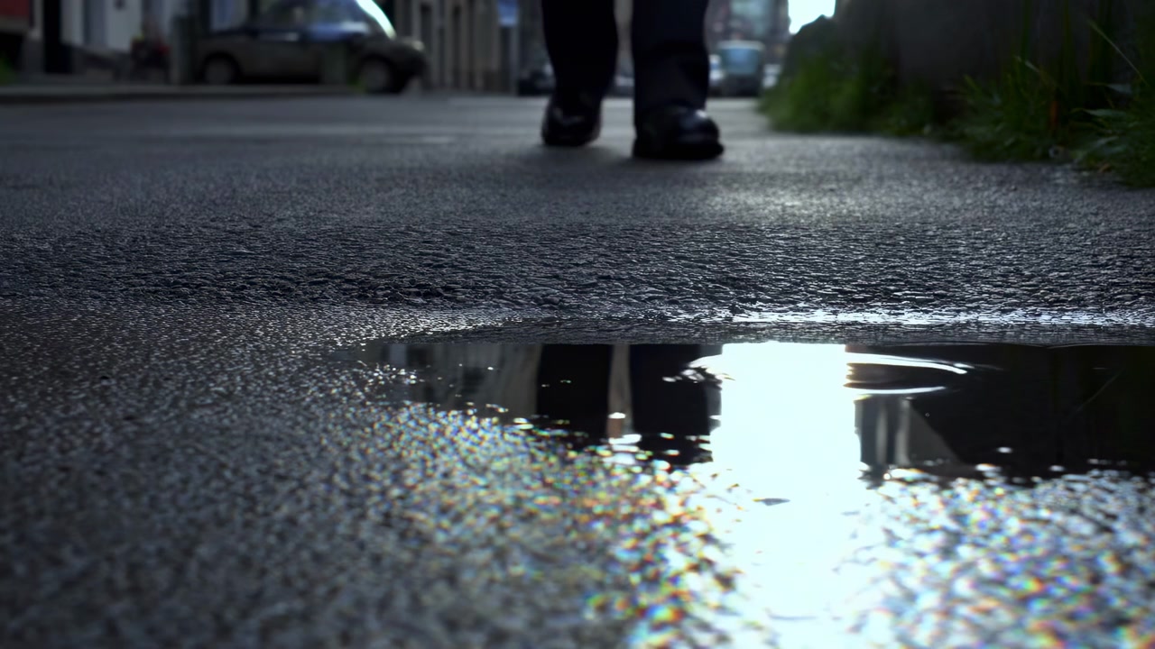 Extreme low-angle urban scene showing wet asphalt, reflective puddle, shallow ripples, blurred pedestrian legs stepping forward, parked cars, soft daylight, and gritty street mood, with puddle