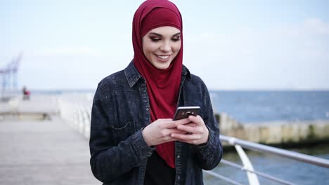 Attraktives-Junges-Mädchen-Mit-Hijab-Auf-Dem-Kopf-Lächelt,-Während-Sie-Jemandem-Eine-SMS-Schreibt-Und-Etwas-Auf-Ihrem-Smartphone-Scrollt.-Zeitlupenaufnahmen-Im-Freien