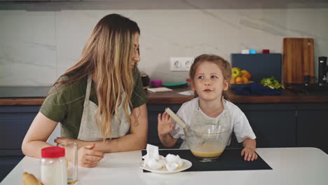 daughter-whisks-egg-in-bowl-with-mother-at-table-in-kitchen