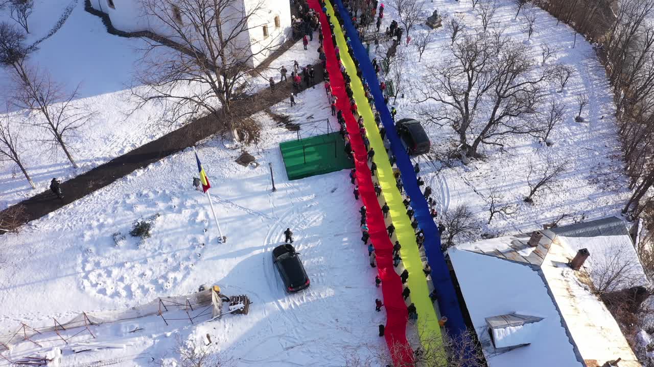 An ascending aerial drone shot of Raducanu Monastery in Targu Ocna, Romania, during winter. A long line of people holds a massive Romanian flag over the snowy grounds in a display of national pride