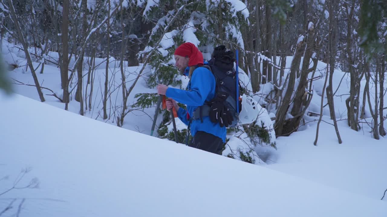 밝은 파란색 재킷과 빨간 모자를 쓴 남자가 깊은 눈 속에서 오르막길을 트레킹하고 있습니다.