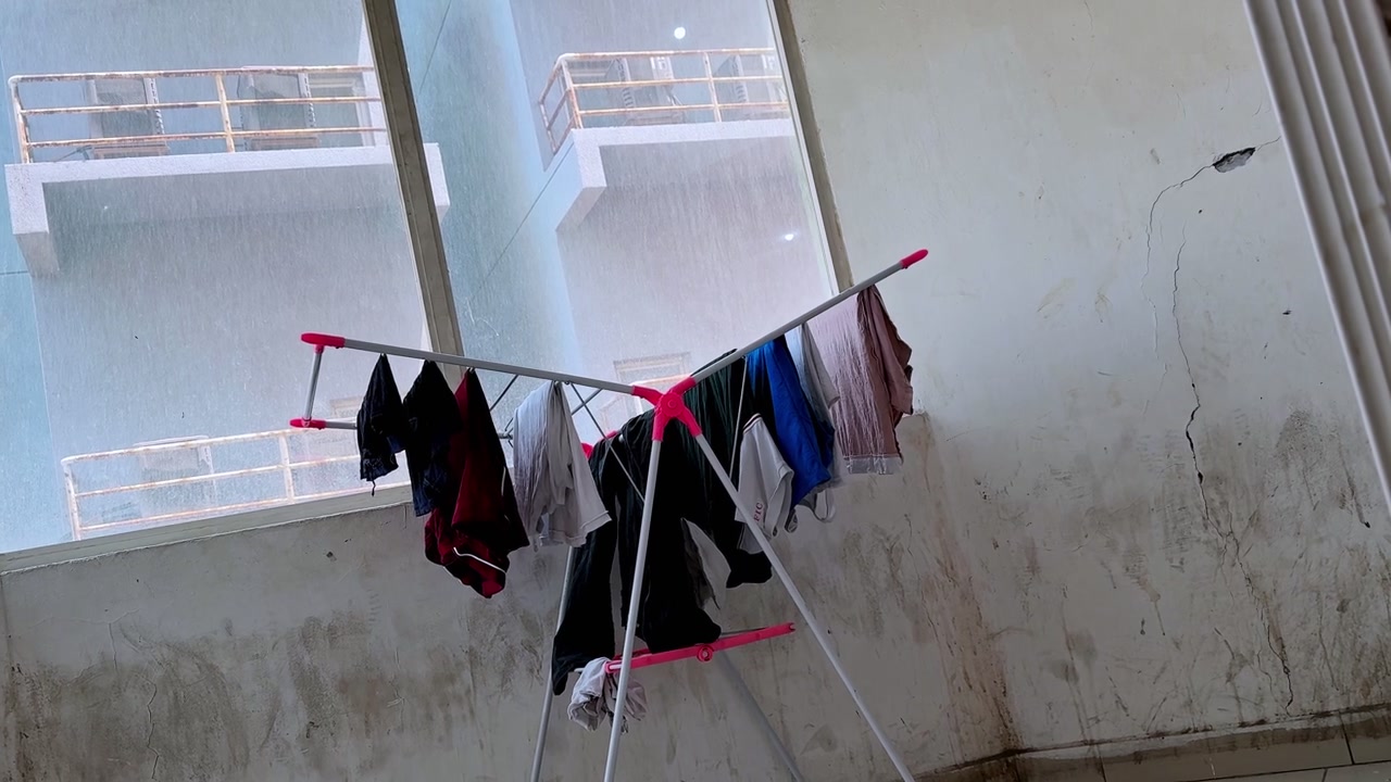 A static shot of laundry hanging on a folding drying rack inside an old apartment during daytime. The room features cracked walls and a large window looking out at neighboring urban buildings.