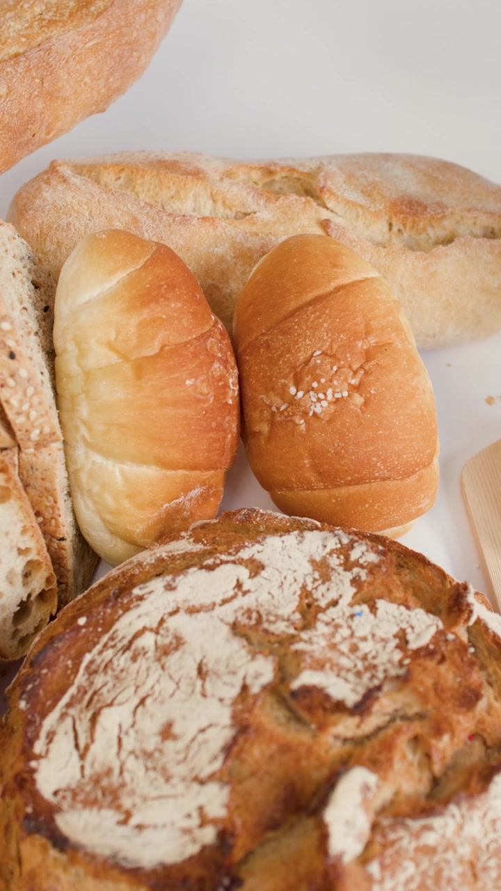 Close-up overhead shot of hands slicing baguette on wooden board surrounded by various breads.