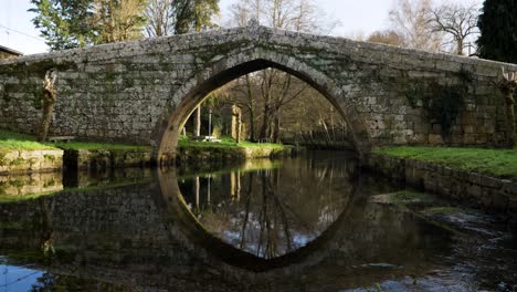 Shallow-water-stream-below-angled-medieval-bridge-with-mossy-and-vines-growing-on-brick