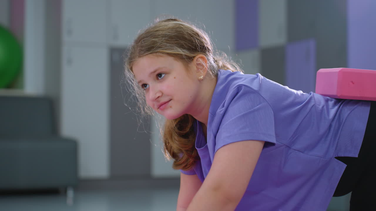 Primo piano ravvicinato di una bambina che estende la mano in avanti durante un esercizio di equilibrio con un blocco viola sulla schiena, enfatizzando la concentrazione e l'equilibrio.