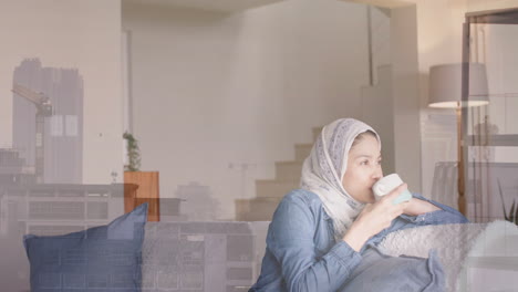 Animation-of-cityscape-over-biracial-woman-in-hijab-drinking-tea