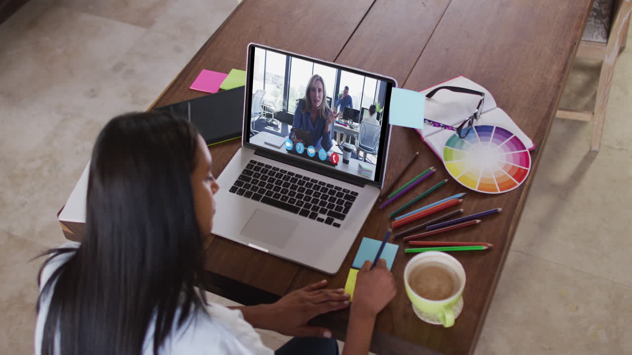 Mujer caucásica usando una laptop en videollamada con una colega, tomando notas