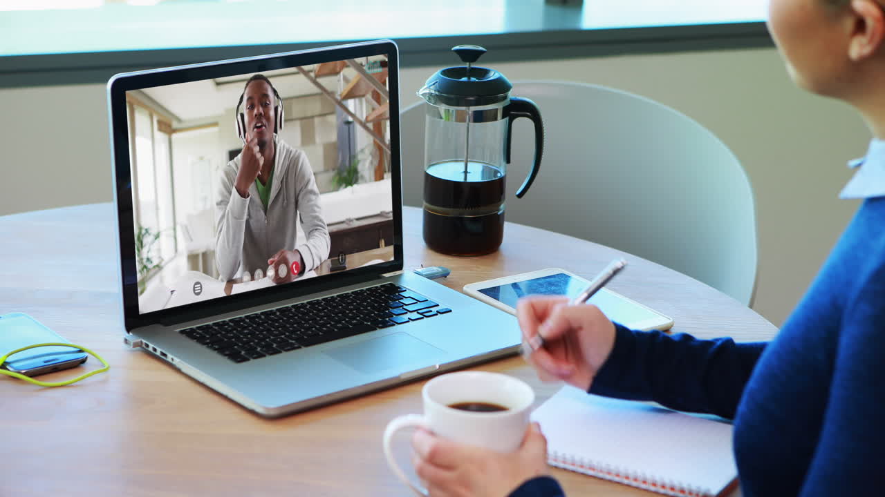 Kaukasisk kvinde, der drikker kaffe og tager noter under et videoopkald på en bærbar computer derhjemme.