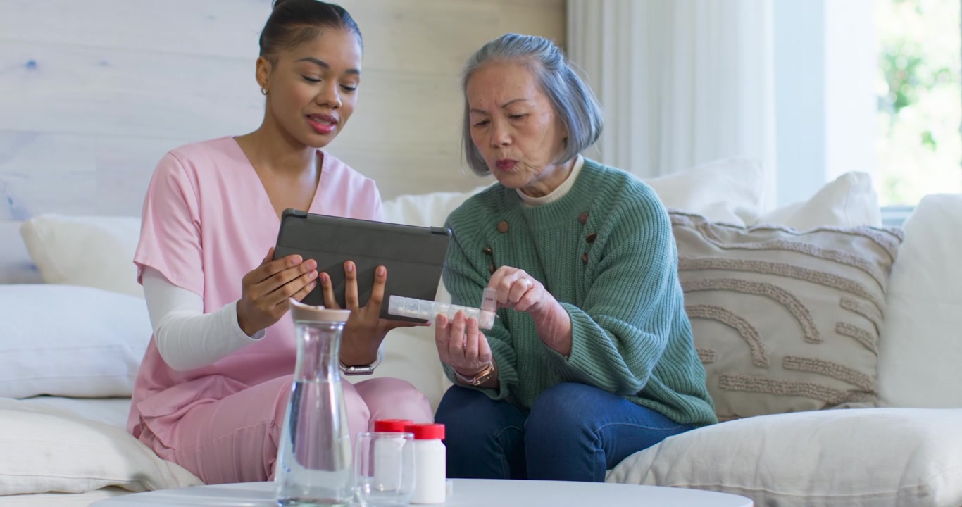 African American caregiver in scrubs guiding Asian senior on sofa using pad confirming blister dose