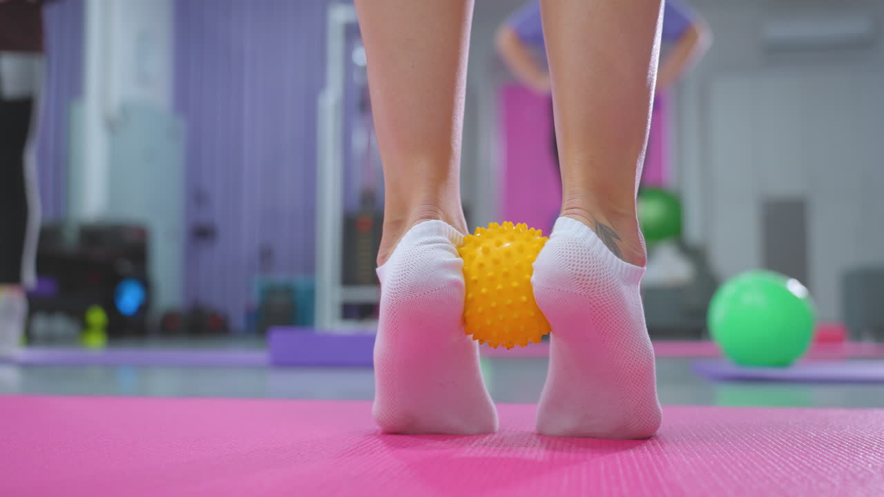 primer plano de una persona con una pierna tatuada, cubierta ligeramente por calcetines, sosteniendo una pelota amarilla puntiaguda bajo el pie para el ejercicio de acumulación de resistencia en una alfombra rosa