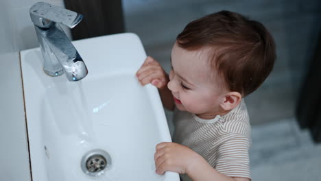 smiling toddler playing with a sink tap. little baby opens and closes the faucet and watches the water flow. high angle view.