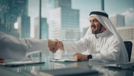 empresario emiratí sonriente en traje tradicional blanco sentado en la oficina con socios de negocios. estrechar la mano en una inversión exitosa. concepto de empresario saudí, emiratí, árabe.