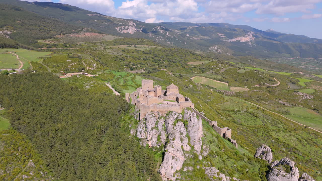 Forward drone shot approaching a medieval stone castle on a rocky hill, surrounded by forest, farmland, and rolling hills under a bright sky. Scenic rural and historic location in northern Spain