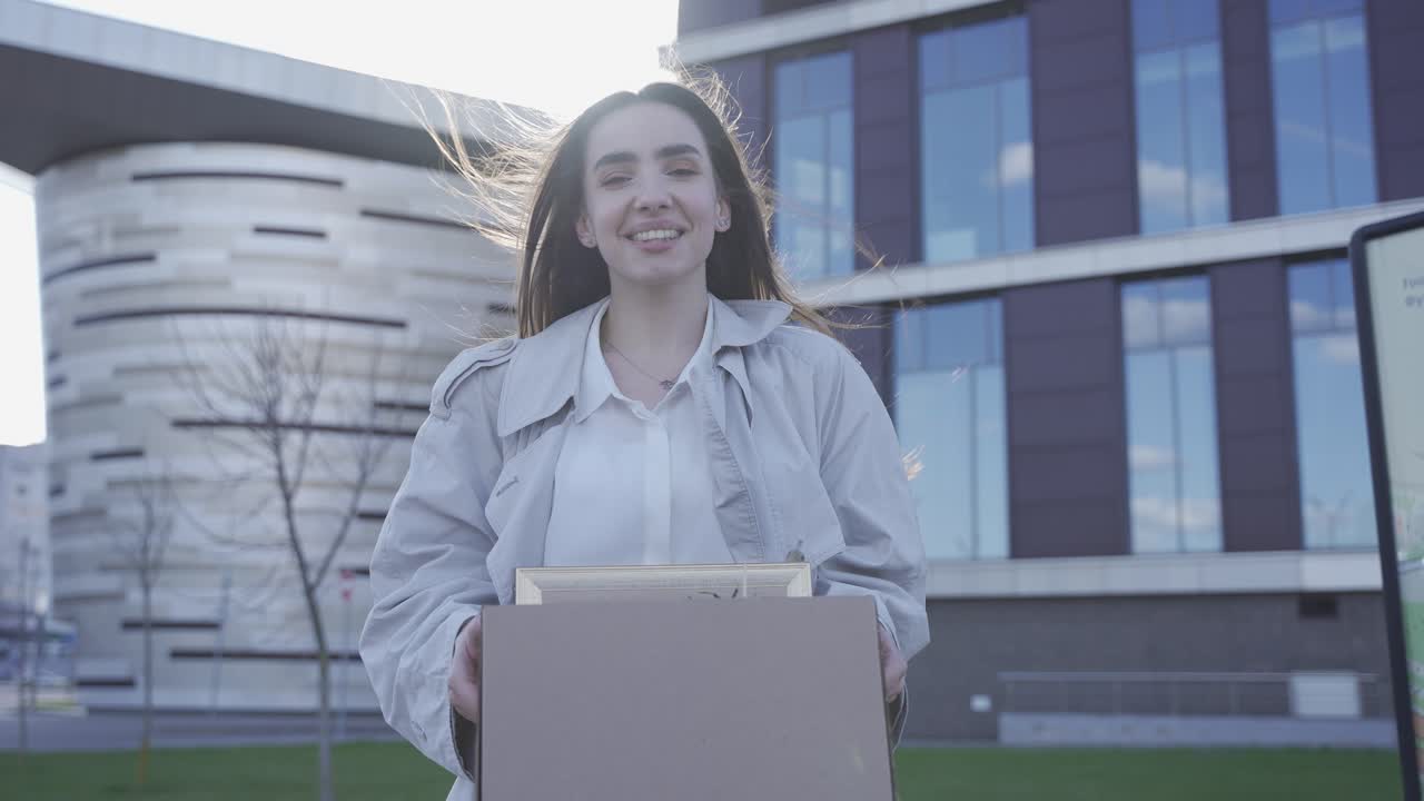 una mujer bonita acaba de dejar su trabajo. ella está frente a un edificio de oficinas. ella es feliz. ella esta sonriendo.