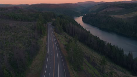 Drohnenaufnahme-Einer-Landstraße-Neben-Einem-Fluss-Bei-Sonnenuntergang-Mit-Fahrenden-Fahrzeugen