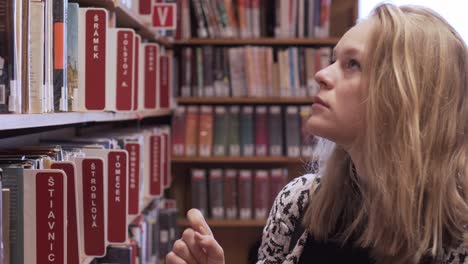 Young-female-is-walking-along-bookshelves,-looking-for-a-right-book-in-library