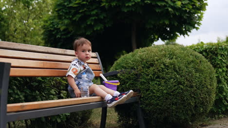 cute caucasian baby boy sits peacefully on the wooden bench. kit shows his tongue playfully. park nature at backdrop.