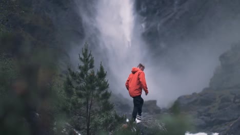 slow motion shot of man walking onto a rock, watching over beautiful scenery, shot taken from behind tree branches, norway mardalsfossen.