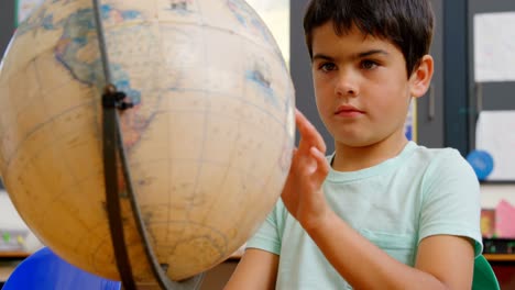 vista frontal de un escolar caucásico estudiando el globo en el escritorio en el aula de la escuela 4k