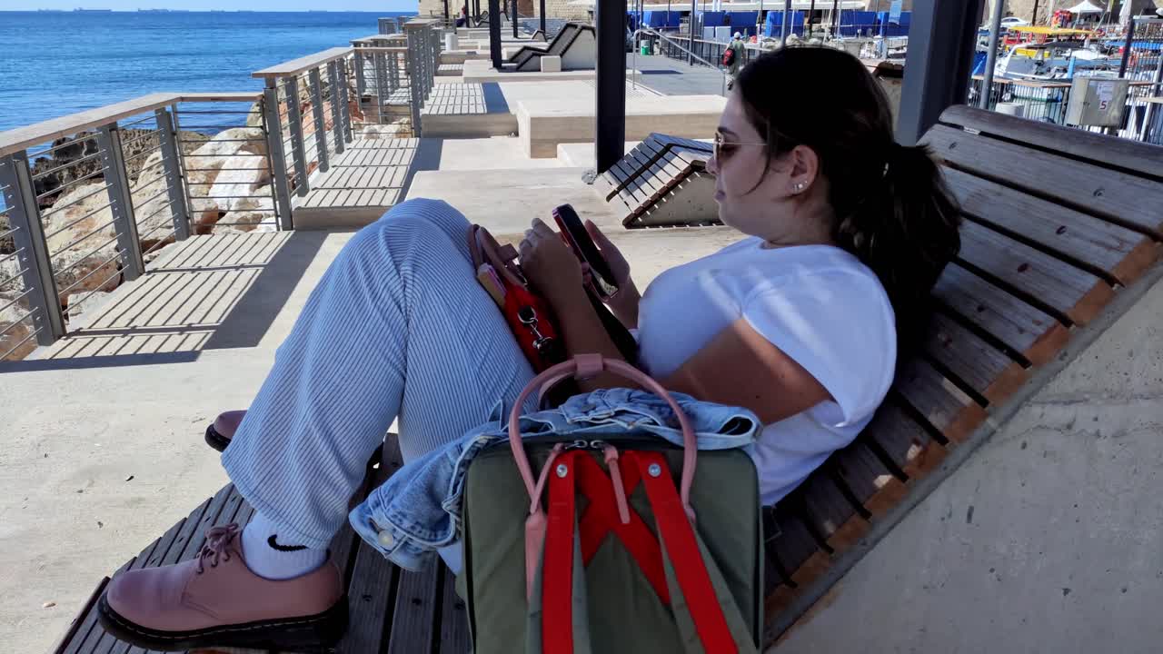 Young woman sitting on a seaside bench with backpack looking at phone