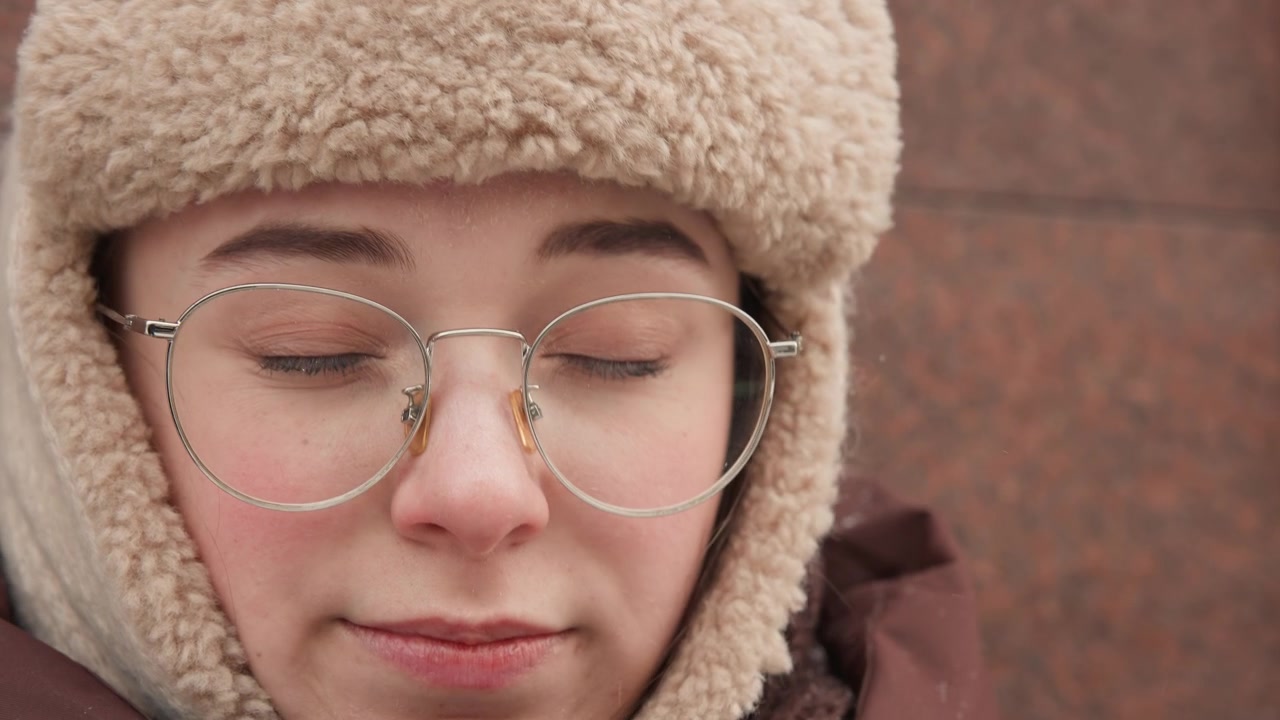 Close-up portrait of young person in shearling-lined hat and hood touching glasses at bridge near forehead, eyes partly closed, expression showing cold discomfort in soft outdoor light