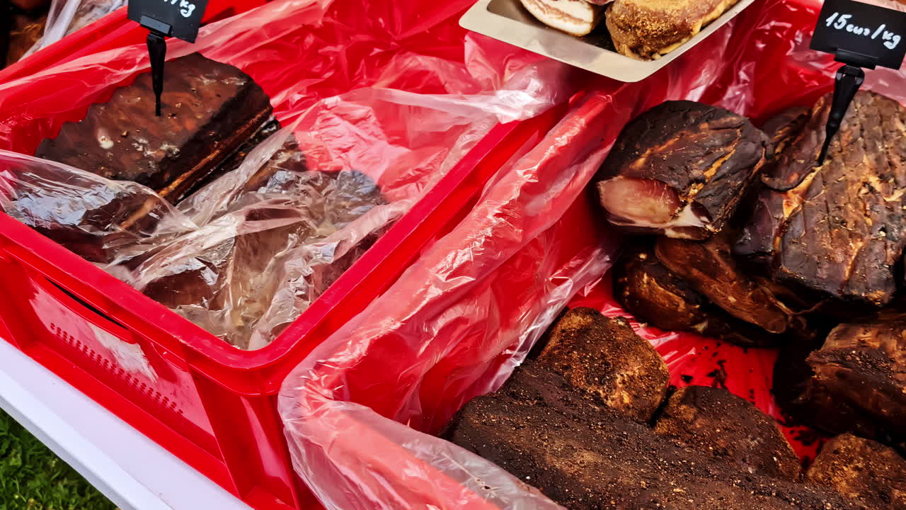 Assorted smoked meats on display in red bins with price tags at a local market stall