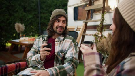 Over-shoulder:-A-happy-brunette-guy-with-a-green-hat-in-a-checkered-shirt-and-his-brunette-girlfriend-drink-tea-from-special-thermos-cups-during-their-picnic-outside-the-city-with-a-trailer