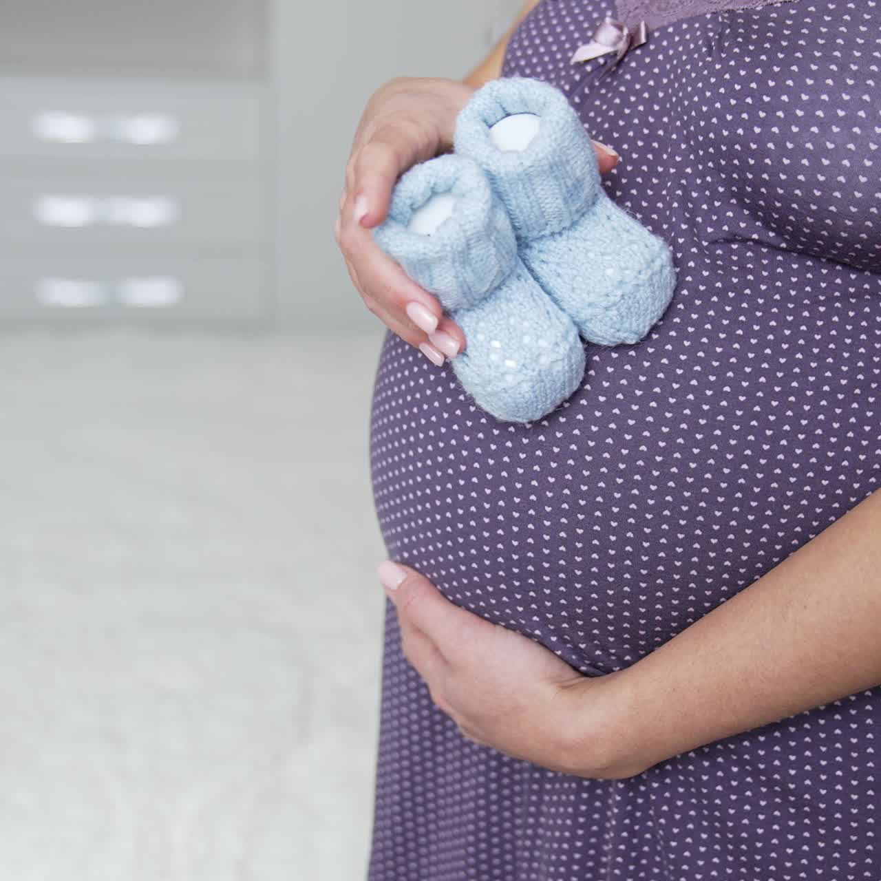 White pregnant woman in a nightgown holding little baby socks with one hand on her belly. She steps with little socks by her stomach. Slow motion