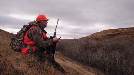 hunter with a rifle sitting on a grassy hill holding a mobile phone searching signal in a hunting field in saskatchewan, canada - medium shot