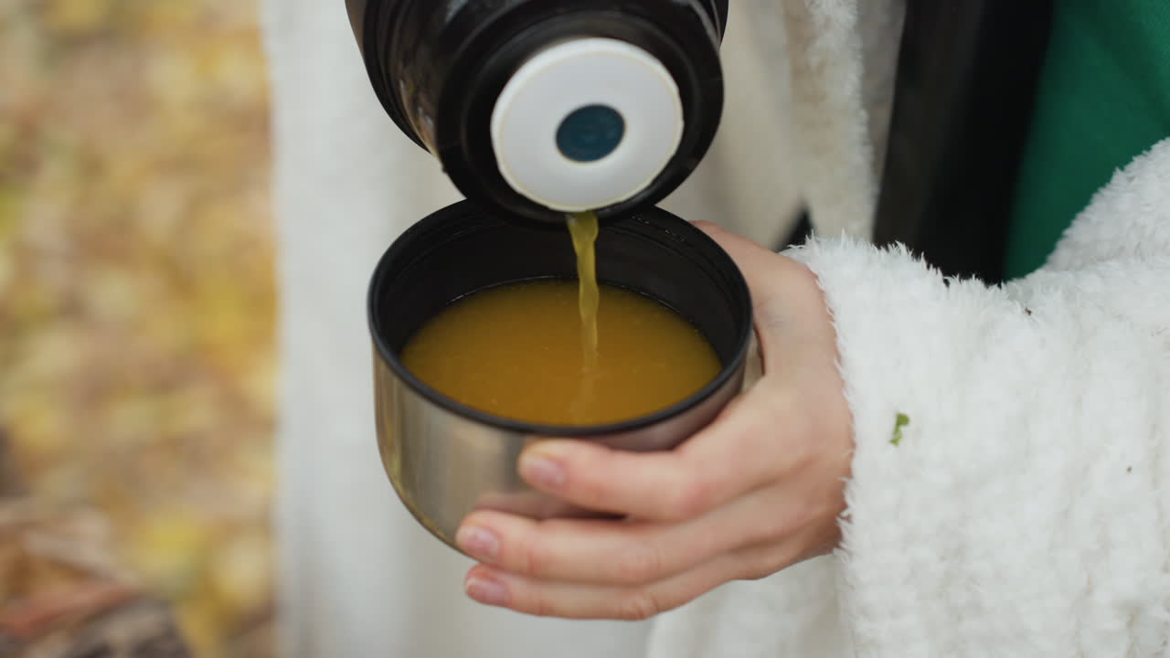 escursionista bianco che versa tè caldo all'aperto, tazza di metallo tra le mani, giacca in pile e maglione verde visibili, foglie cadute che ricoprono il terreno, vapore che sale dal liquido ambrato, versamento lento nella tazza in acciaio inossidabile