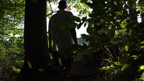 young man walks barefoot in slow motion out of a dark forrest towards a bright green meadow in with sunshine