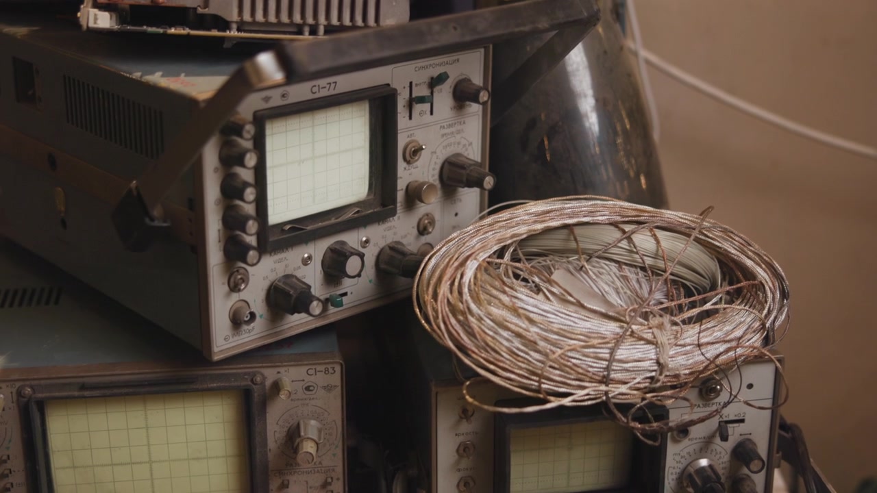 Close view of stacked vintage oscilloscopes resting on workshop shelf with braided copper wire, showing CRT screens, knobs, dials, dust, patina, reflections, and retro industrial detail