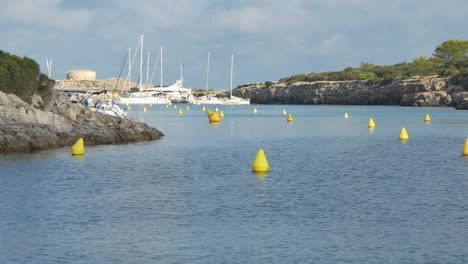 Calm-view-at-sunrise-of-creek-Cala-Santandria-in-Menorca-with-yellow-buoys-and-boats,-blue-sea-and-surrounding-rocks