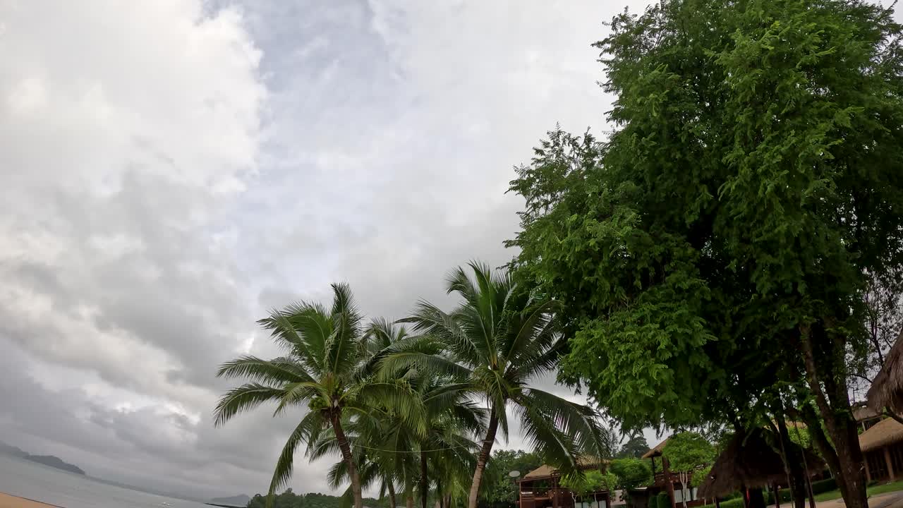 Wide-angle camera pans slowly along a quiet sandy beach with coconut palms, hammock, and resort buildings under overcast skies at a Thai island location