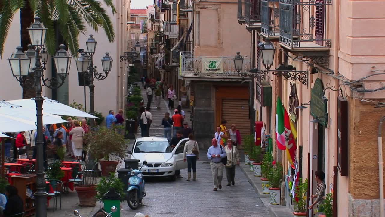 Pedestres passam por luminárias e edifícios de tijolos muito próximos uns dos outros em Cefalù, Itália.