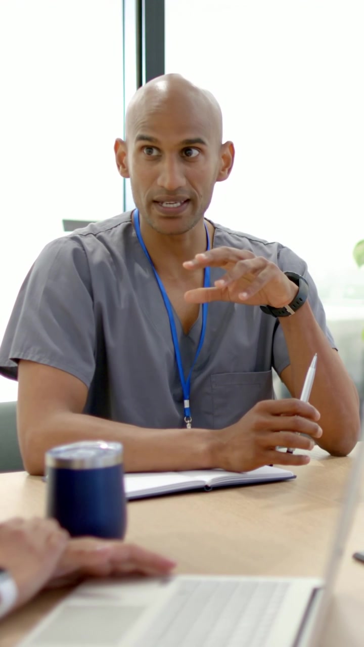Vertical video: Gesturing doctor in grey scrubs explaining case mid talk, holding pen with laptop