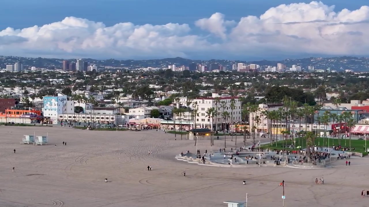 En 70 mm drönarbild tagen från låg höjd med framåtroterande kamera över Venice Beach Skate Park vid solnedgången längs Ocean Front Walk, med folkmassor av skejtare och rosa-lila himmel över Los Angeles, Kalifornien