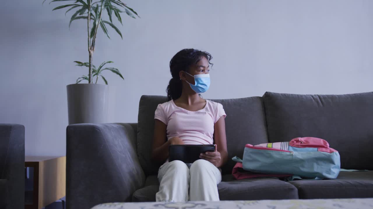 Afroamerikansk jente med munnbind bruker nettbrett mens hun sitter i sofaen på sykehuset.