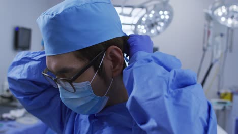 asian male surgeon wearing face mask, putting surgical cap on in operating theatre