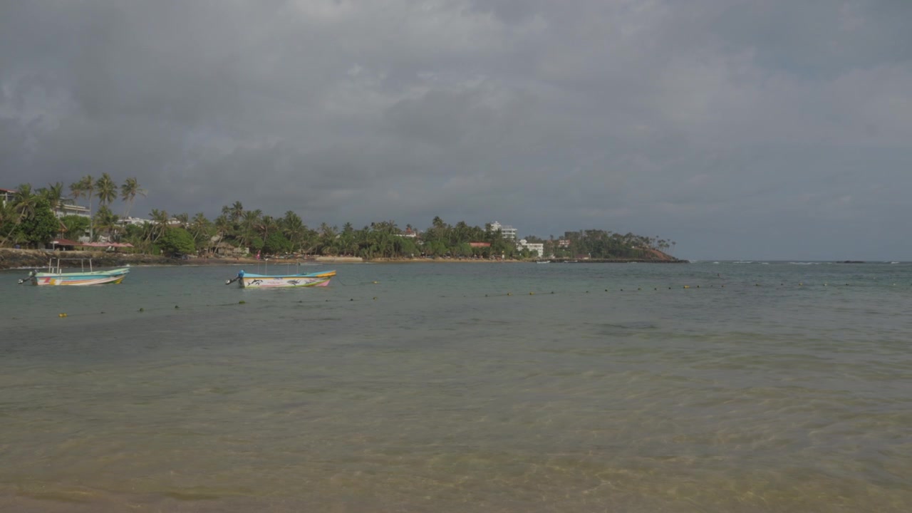 Una piccola barca a motore attraversa una baia tropicale fosca davanti a una costa fiancheggiata da palme, creando una debole scia in lontananza vicino a Mirissa, Sri Lanka
