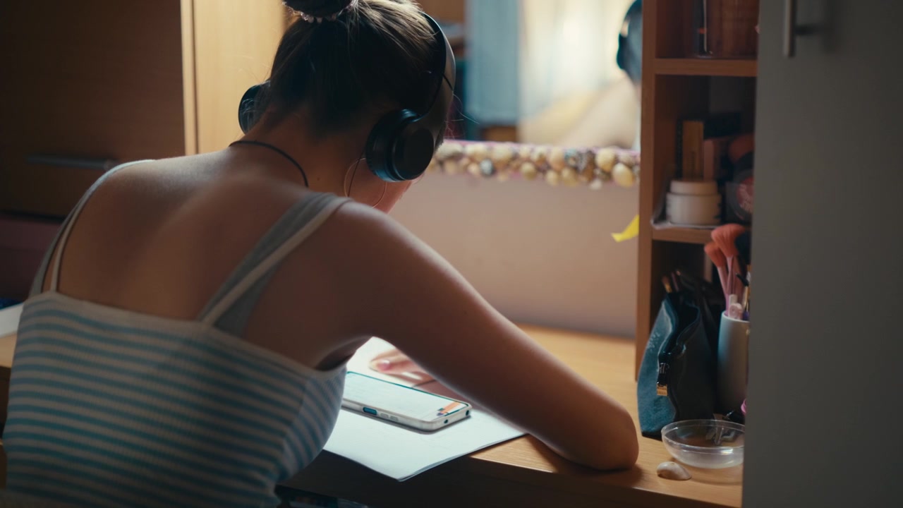 Rear view of a young female student sitting at her desk, wearing over-ear headphones and listening to music while trying to focus on textbook notes. Retro analog cinematic film look, 4k.
