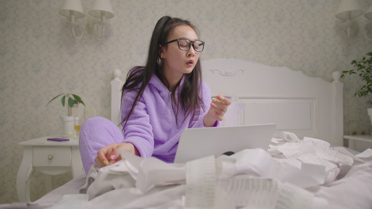 안경을 쓴 아시아 여성이 침대에 앉아 상점 영수증을 보고 있습니다. 20대 여성은 노트북을 사용하여 가족 예산을 회계합니다.