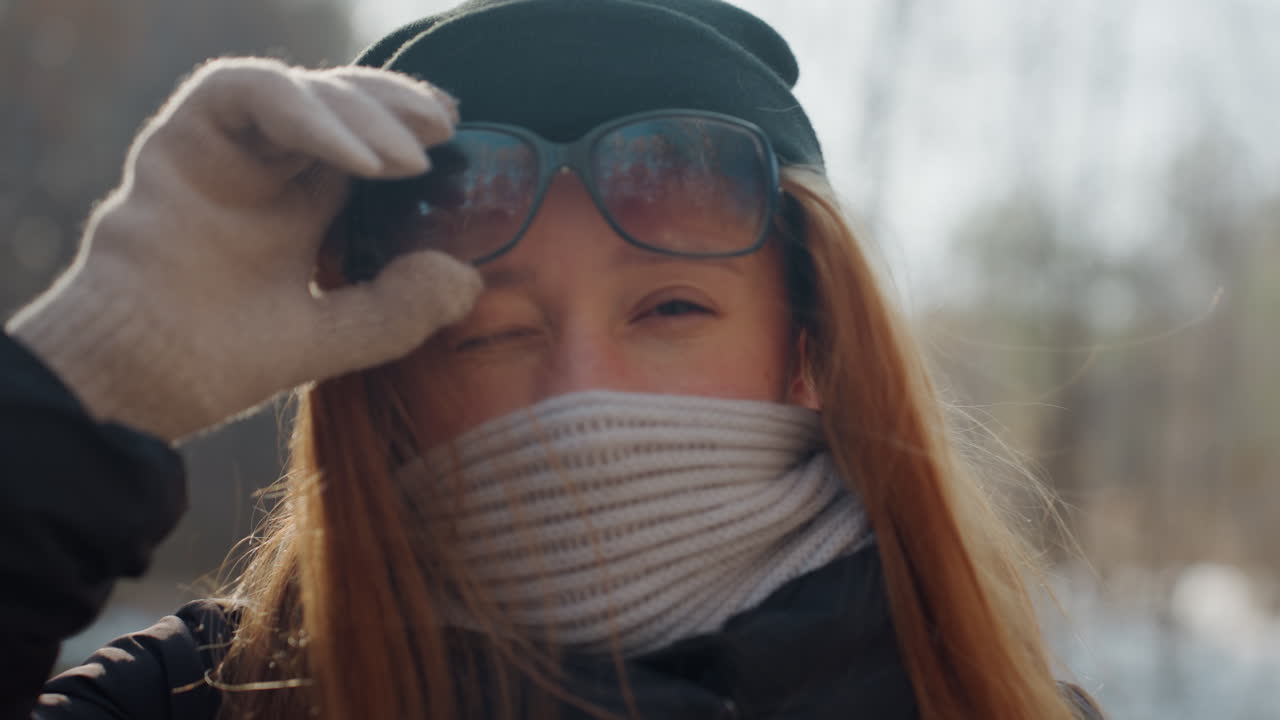 Bílá žena si v zasněženém lese sundává masku, rukou v rukavici si zvedá sluneční brýle, aby odhalila oči, šála a kulich, jemné sluneční světlo a protisvětlo, výraz se mění z ostražitého na uvolněný, cestovatelka