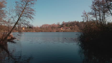 drone flying over tranquil lake immersed in the woods overlooking countryside landscape during sunset in italy - aerial drone shot